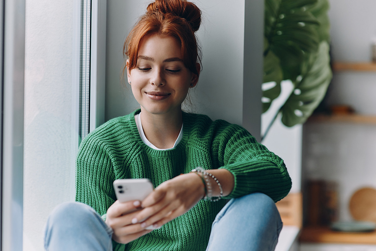 attractive-young-woman-using-smart-phone-smiling-while-sitting-window-sill-home
